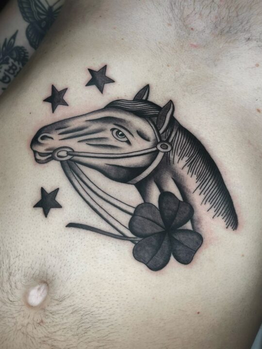 Cabeza de caballo con brida junto a un trébol de cuatro hojas y tres estrellas, tatuada en el pecho en negro y gris.