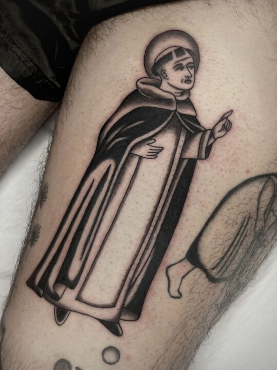 Fraile con hábito y aureola señalando hacia arriba con el índice, tatuado en el muslo en negro y gris.