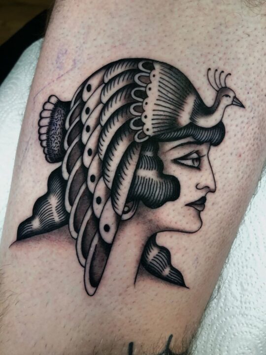 Perfil de mujer con un pavo real desplegado sobre la cabeza a modo de tocado, tatuado en el brazo en negro y gris.