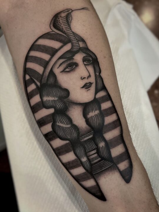 Retrato de mujer con tocado egipcio de rayas y una cobra erguida en la frente, tatuado en el antebrazo en negro y gris.