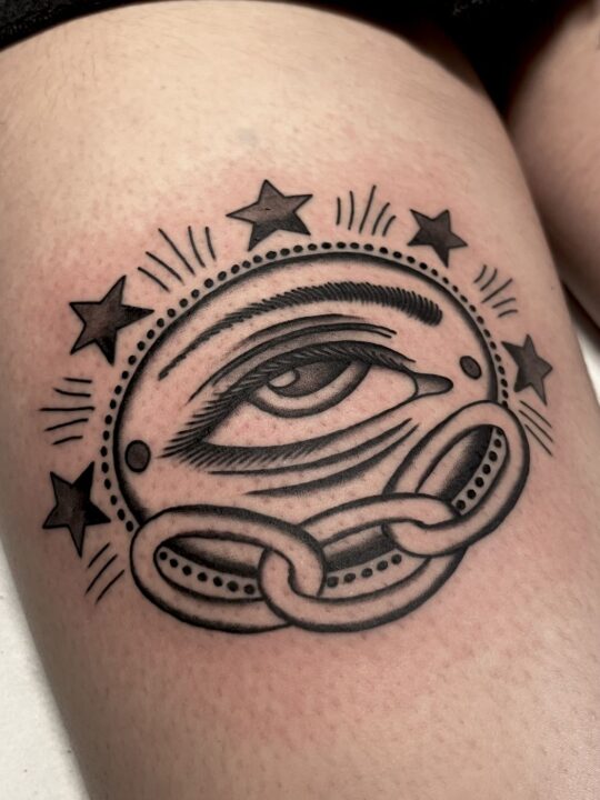 Ojo dentro de un disco perlado, rodeado de cinco estrellas y cruzado por eslabones de cadena, tatuado en el muslo.