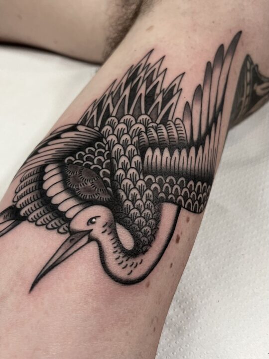 Grulla en vuelo con las alas desplegadas y el plumaje escamado, tatuada en la cara interna del brazo.