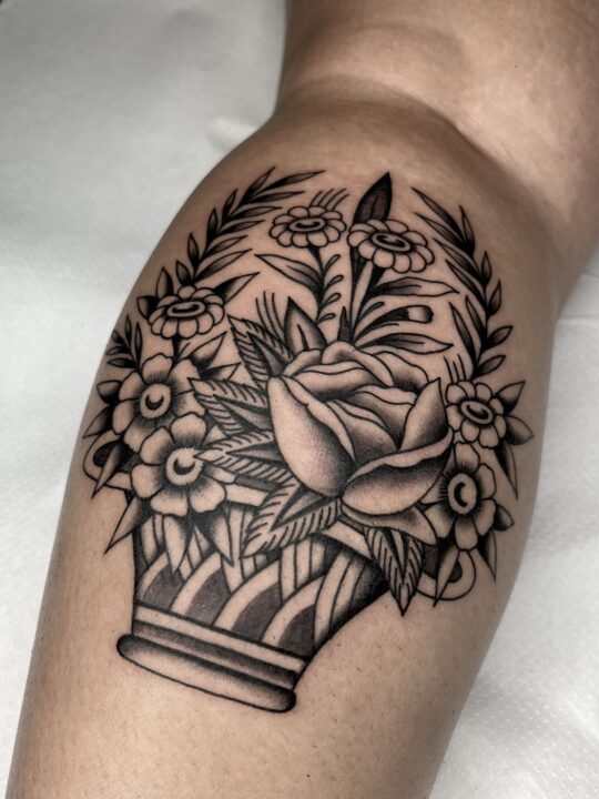 Cesto rayado lleno de flores con una rosa en el centro, tatuado en la pantorrilla en negro y gris.
