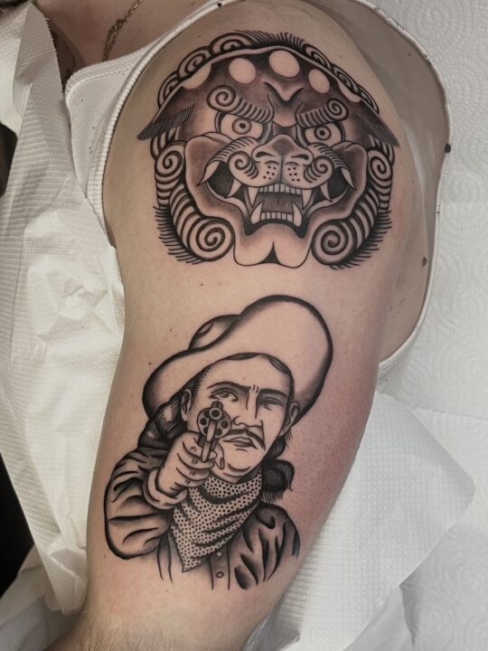 Máscara de león shishi con volutas y, debajo, un vaquero con sombrero apuntando con un revólver, tatuados en el brazo.