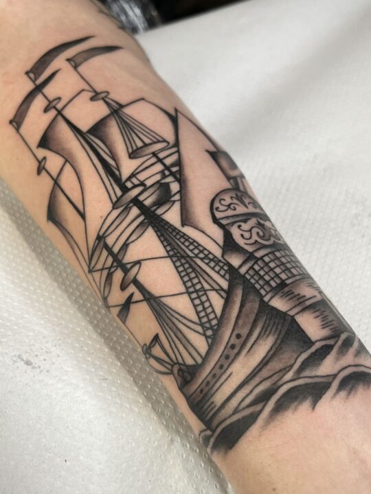 Velero de tres mástiles con las velas henchidas sobre las olas, tatuado en el antebrazo en negro y gris.
