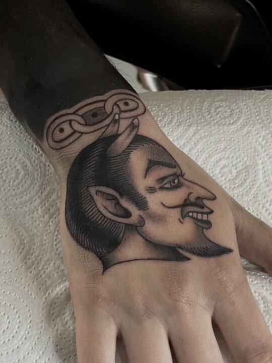 Cabeza de diablo con cuernos y perilla, con eslabones de cadena en la muñeca, tatuada en el dorso de la mano.