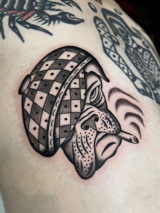Bulldog con gorra de cuadros fumando un cigarrillo, tatuado en el costado en negro y gris.