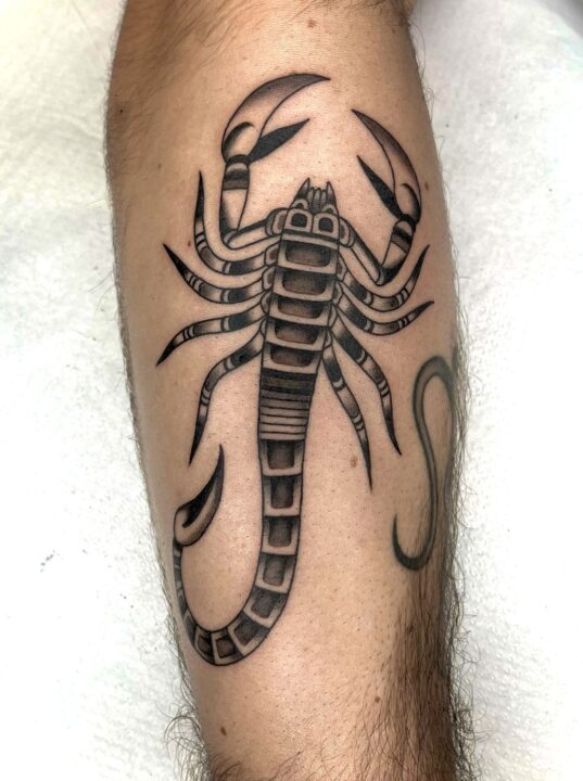 Escorpión con las pinzas abiertas y la cola levantada, tatuado en la pierna en negro y gris.