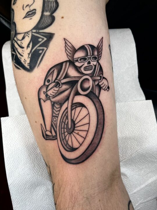 Calavera con casco alado y gafas montada en una moto, tatuada en la pierna en negro y gris.