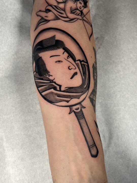 Espejo de mano con el rostro de un actor de kabuki reflejado, tatuado en el antebrazo en negro y gris.