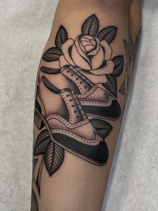 Par de zapatos de vestir picados junto a una rosa con hojas, tatuados en la pierna en negro y gris.