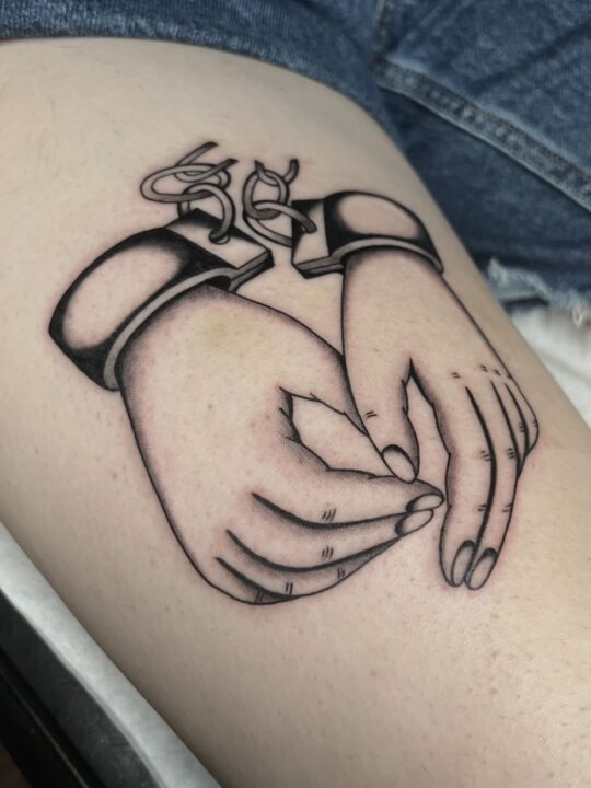 Dos manos cruzadas y esposadas por las muñecas, tatuadas en el muslo en línea negra.