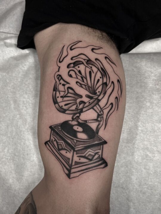 Gramófono de trompa con el sonido saliendo en volutas, tatuado en el muslo en negro y gris.
