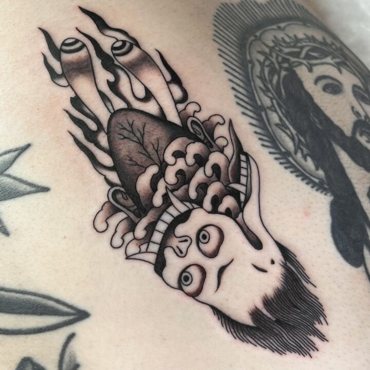 Cabeza cortada de estilo japonés envuelta en llamas y olas, tatuada en el costado en negro y gris.