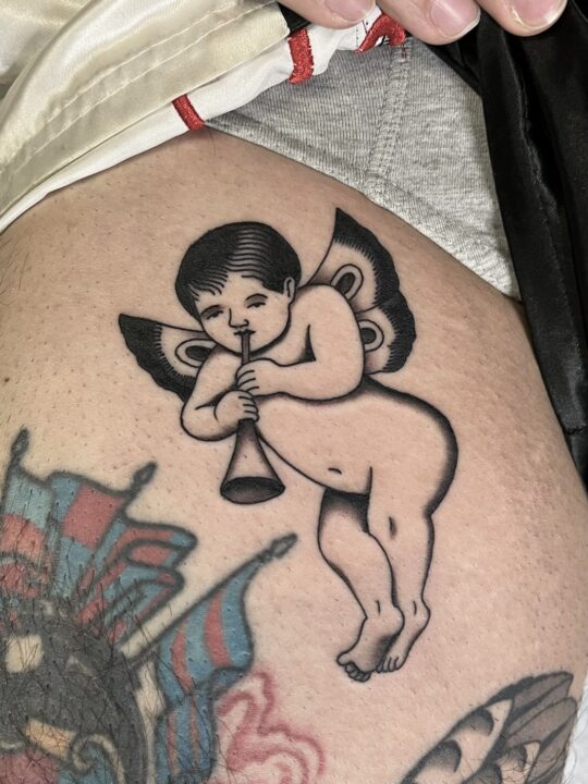 Querubín alado tocando una trompeta, tatuado en la cadera en negro y gris.