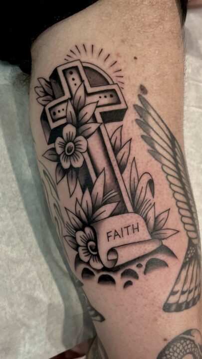 Cruz con rayos y flores sobre una filacteria con la palabra FAITH, tatuada en el muslo.