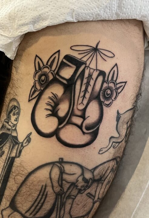 Par de guantes de boxeo colgados entre flores, tatuados en el muslo en negro y gris.
