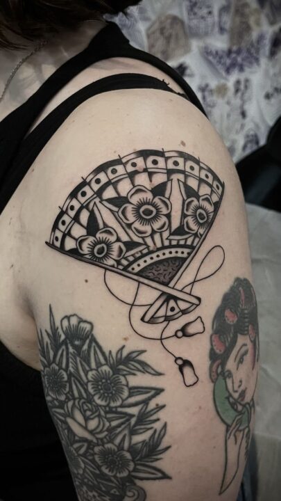 Abanico abierto decorado con flores y con la borla colgando, tatuado en el hombro en negro y gris.