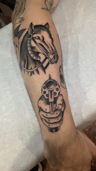 Cabeza de caballo con brida y, debajo, una mano apuntando con un revólver, tatuados en el antebrazo.