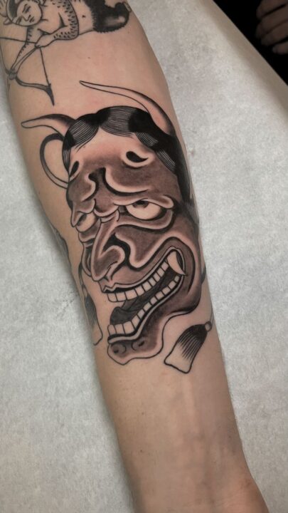 Máscara hannya de cuernos largos y dientes apretados, tatuada en el antebrazo en negro y gris.
