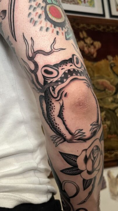 Rana con astas de ciervo abrazada al codo, tatuada en el brazo en negro y gris.