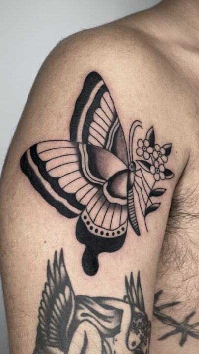 Mariposa de alas negras junto a un ramillete de flores pequeñas, tatuada en el hombro.