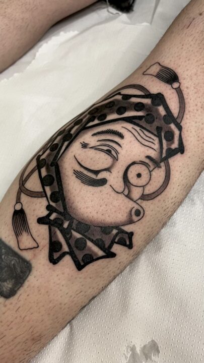 Rostro guiñando un ojo con monóculo, pañuelo de lunares en la cabeza y cuello de arlequín, tatuado en la pierna.