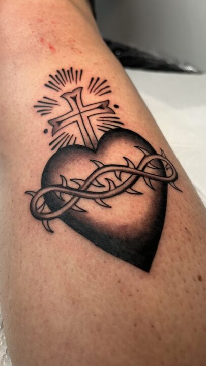 Corazón negro atravesado por una corona de espinas con una cruz radiante encima, tatuado en el muslo.
