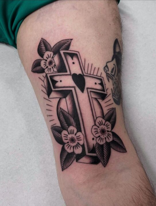 Cruz con un corazón en el centro rodeada de flores y hojas, tatuada en el muslo en negro y gris.