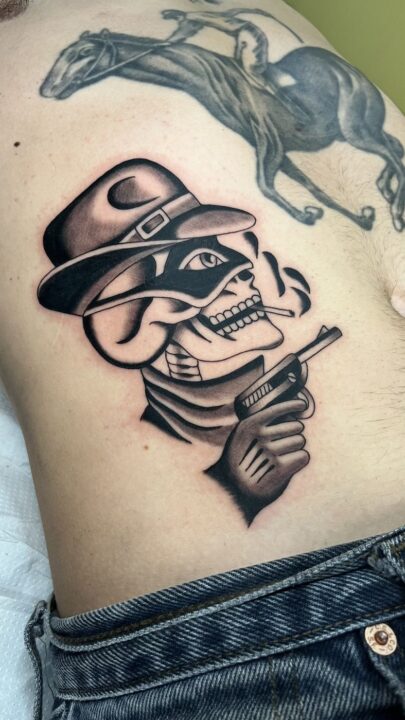 Calavera con sombrero de ala ancha, cigarrillo y revólver en la mano, tatuada en el costado.