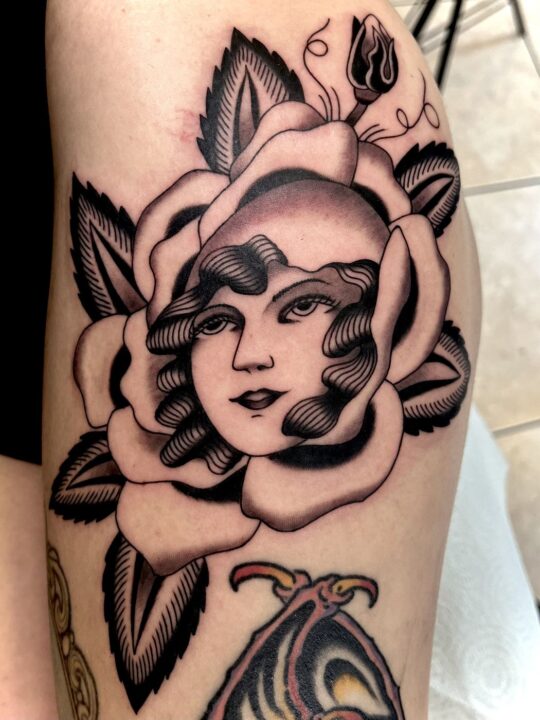 Rostro de mujer con el pelo ondulado dentro de una rosa abierta, tatuado en el muslo.