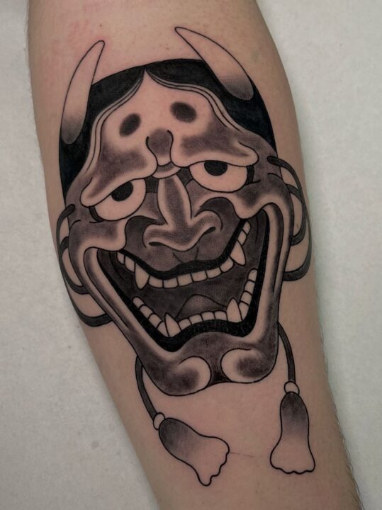 Máscara hannya con cuernos y borlas colgando, tatuada en la pierna en negro y gris.