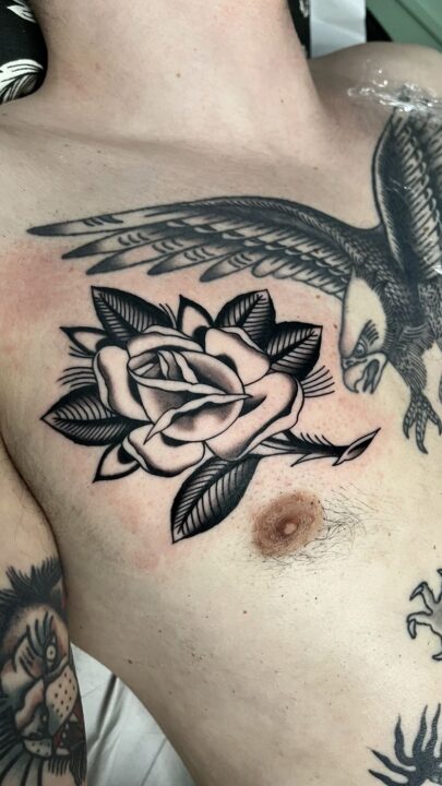 Rosa abierta con hojas bajo un águila con las alas desplegadas, tatuadas en el pecho.