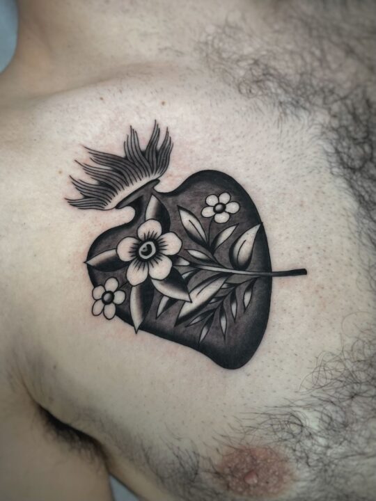 Corazón negro en llamas atravesado por una rama con flores, tatuado en el pecho.
