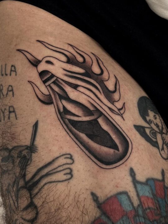 Forma alargada envuelta en llamas con dos ganchos en la punta, tatuada en la cadera en negro y gris.