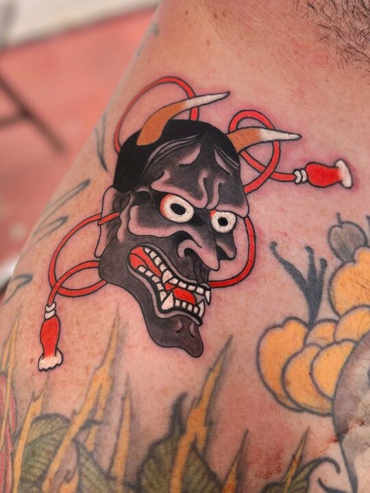 Máscara hannya gris con la boca roja y un cordón rojo con borlas alrededor, tatuada en el hombro.