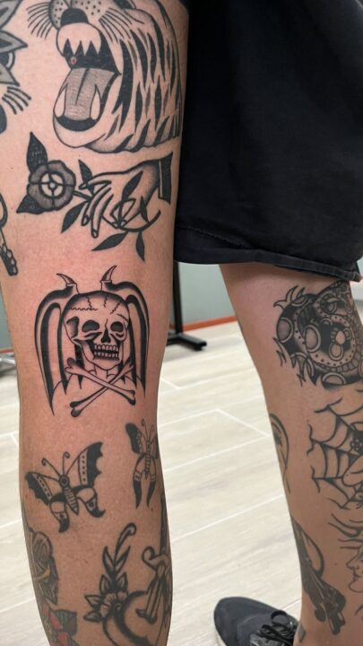 Pierna cubierta de tatuajes con una calavera de alas de murciélago sobre dos huesos cruzados en el centro.