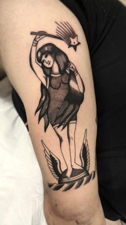 Mujer con una vara en alto de pie sobre una rueda alada, con una estrella fugaz al fondo, tatuada en el brazo.