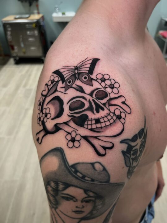 Calavera sonriente sobre huesos cruzados, con una mariposa encima y flores alrededor, tatuada en el hombro.