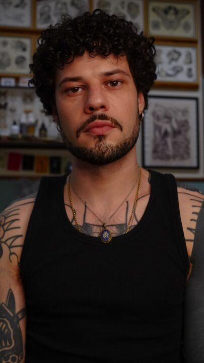 Retrato de Augusto Cabrera en el estudio, con camiseta de tirantes negra, delante de la pared de láminas enmarcadas.